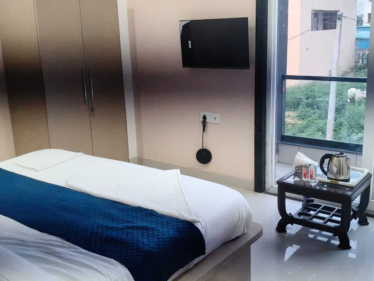 A hotel room with a bed, a TV mounted on the wall, a small table with a kettle, and a window with a view of buildings.
