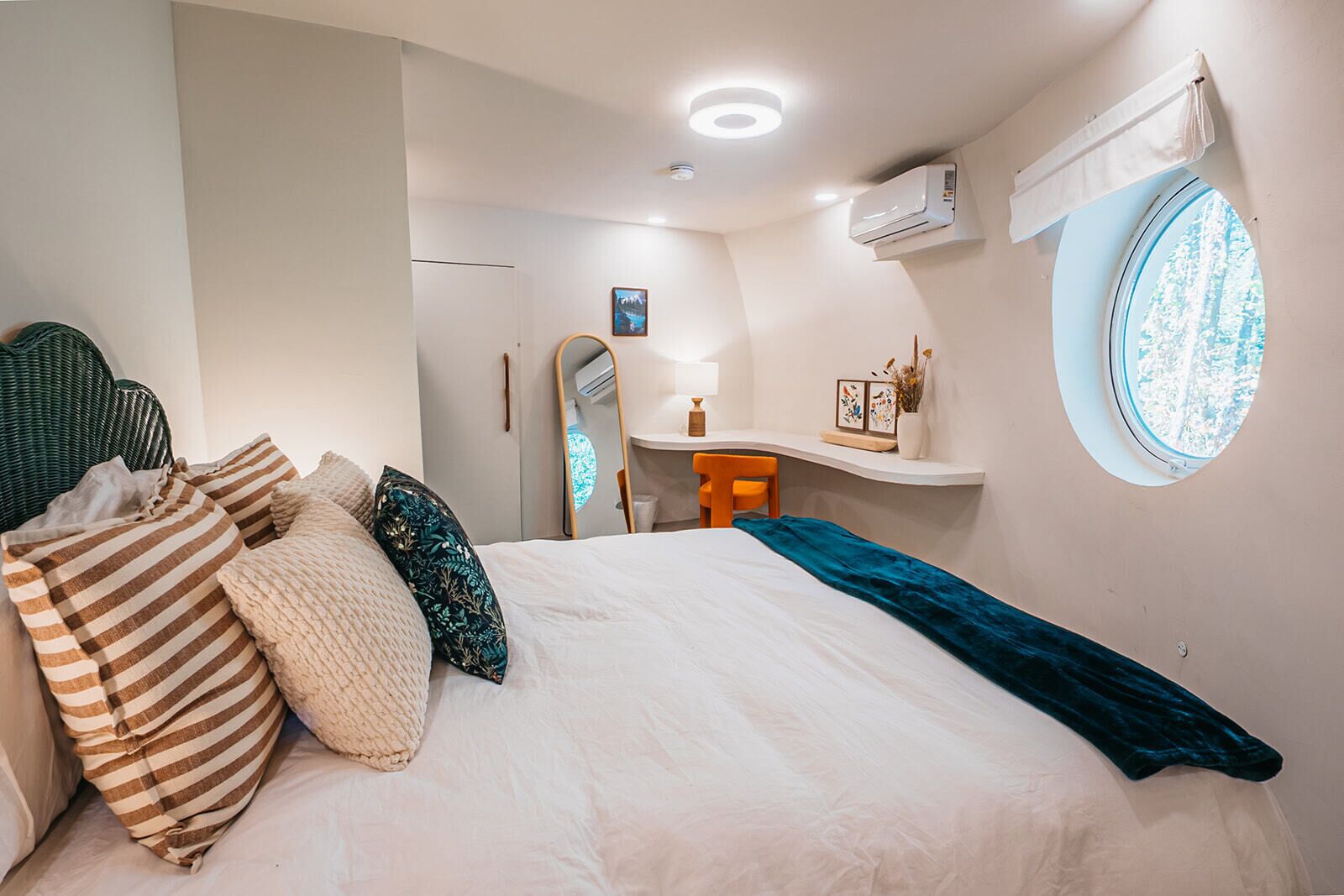 A bedroom with a bed, a desk, a chair, a mirror, and a window.