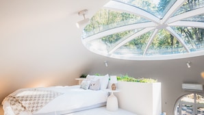 A modern bedroom with a curved ceiling and a skylight, a bed with white bedding, and a small bedside table with a lamp.