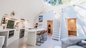 A modern kitchen with a central island, a dining area with chairs, a staircase, and a cozy seating area with a white fur throw.