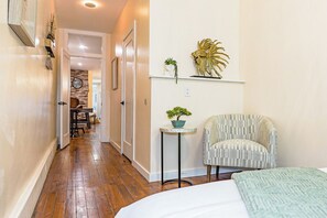 A hallway with wooden flooring leading to a room with a bed, a chair, and a small table with a plant. - Luxury 3BR/3BA•Air Hockey Lounge•Walkable Old City (Philadelphia)