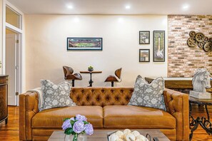 Ein Wohnzimmer mit einem braunen Ledersofa, zwei Sesseln, einem Couchtisch mit Blumen und einer Backsteinwand mit Dekorationselementen.
