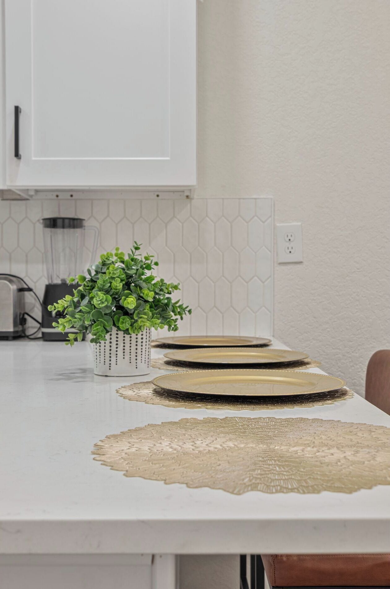 A kitchen with white cabinets, a hexagonal backsplash, a blender, a plant in a white pot, and gold-rimmed plates on a table.