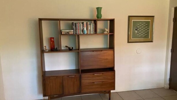 Een houten boekenkast met meerdere planken en lades, een groene vaas bovenop en een ingelijst diploma aan de muur.