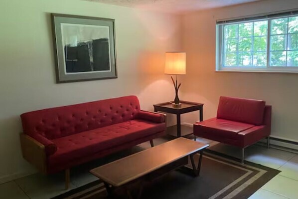 Ein Zimmer mit einem roten Sofa, einem Holztisch, einem kleinen Beistelltisch mit Lampe und einem Fenster mit Blick auf Bäume.