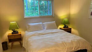 Een slaapkamer met een bed, twee nachtkastjes met lampen, een raam met uitzicht op groen, en een klein afvalbakje.