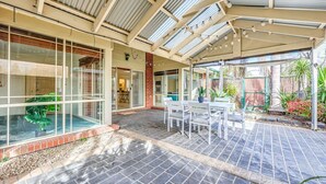 A covered patio area with a table and chairs, a glass enclosure with plants, and a brick wall. - StayAU Family Retreat w/Pool Table in Cranbourne (Cranbourne)