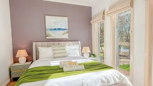 A bedroom with a large bed, two bedside tables with lamps, a painting on the wall, and a view of the outdoors through a window.