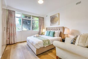 Ein Schlafzimmer mit Bett, Nachttischlampen, Kopfteil, einem Fenster mit Vorhängen und einem Wandbild.