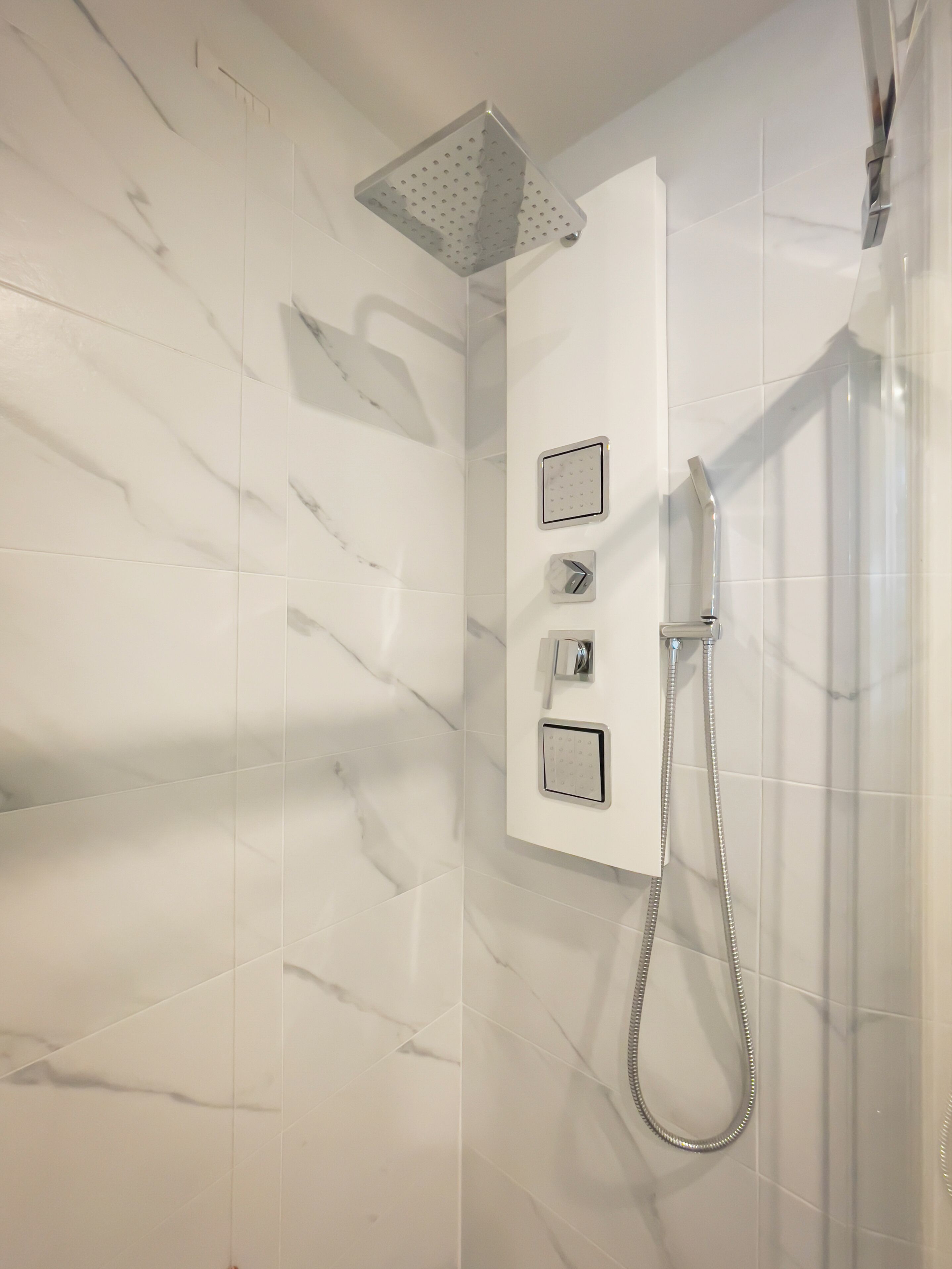 A modern bathroom with white marble tiles, a wall-mounted shower with a handheld showerhead, and two control panels.