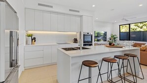 Una cocina moderna con armarios blancos, una isla central con taburetes, horno empotrado y un amplio ventanal con vistas a la vegetación.