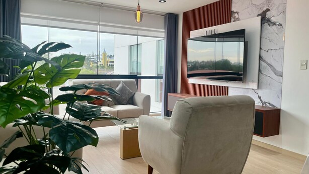 A modern living room with a sofa, a TV mounted on the wall, and large windows offering a city view. - Modern apartment Vallecito (Arequipa)