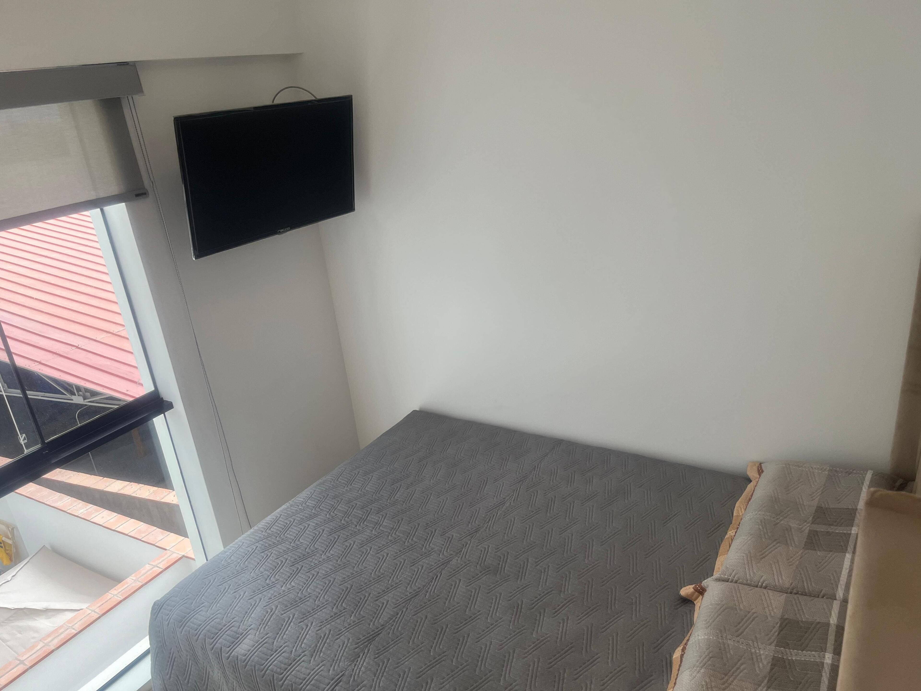 A small, compact room with a bed, a television mounted on the wall, and a window providing a view of the outside.