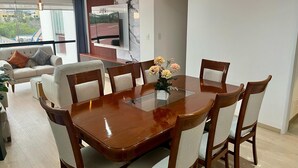 A modern dining area with a wooden table and chairs, a vase of flowers, and a large window with a view of buildings. - Modern apartment Vallecito (Arequipa)