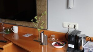 A hotel room with a wooden desk, a television, a coffee maker, a glass of water, a mug, a small plant, and a bowl of fruit. - Lachnerhof (Erlangen)