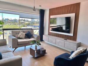 A modern living room with a large TV, a sofa, a coffee table, and a view of the city through large windows.