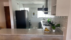 A modern kitchen with a granite countertop, a refrigerator, a stove, and a sink. There are three white bar stools in front of the counter.