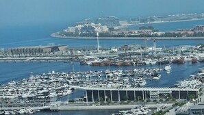 Una marina con numerosas embarcaciones amarradas, un edificio moderno con un diseño de techo singular, y una vista distante de un paisaje urbano costero con edificios y otra marina.