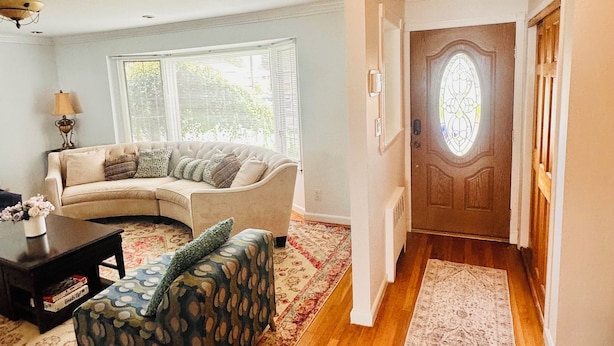 A living room with a sofa, coffee table, and patterned rug. A hallway leads to a door with a stained glass window. - Spacious 5BR Home Private Backyard Basket Ball Hoop Near NY& Long Island Beaches (Syosset)