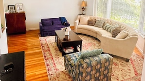 A living room with a sofa, a coffee table, and a patterned ottoman. - Spacious 5BR Home Private Backyard Basket Ball Hoop Near NY& Long Island Beaches (Syosset)