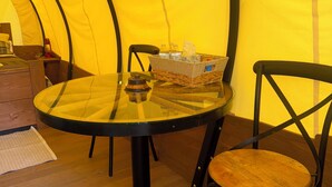 A round glass table with a wicker basket containing bottled water and a tissue, a black metal chair with a wooden seat, and a yellow tent-like structure in the background.