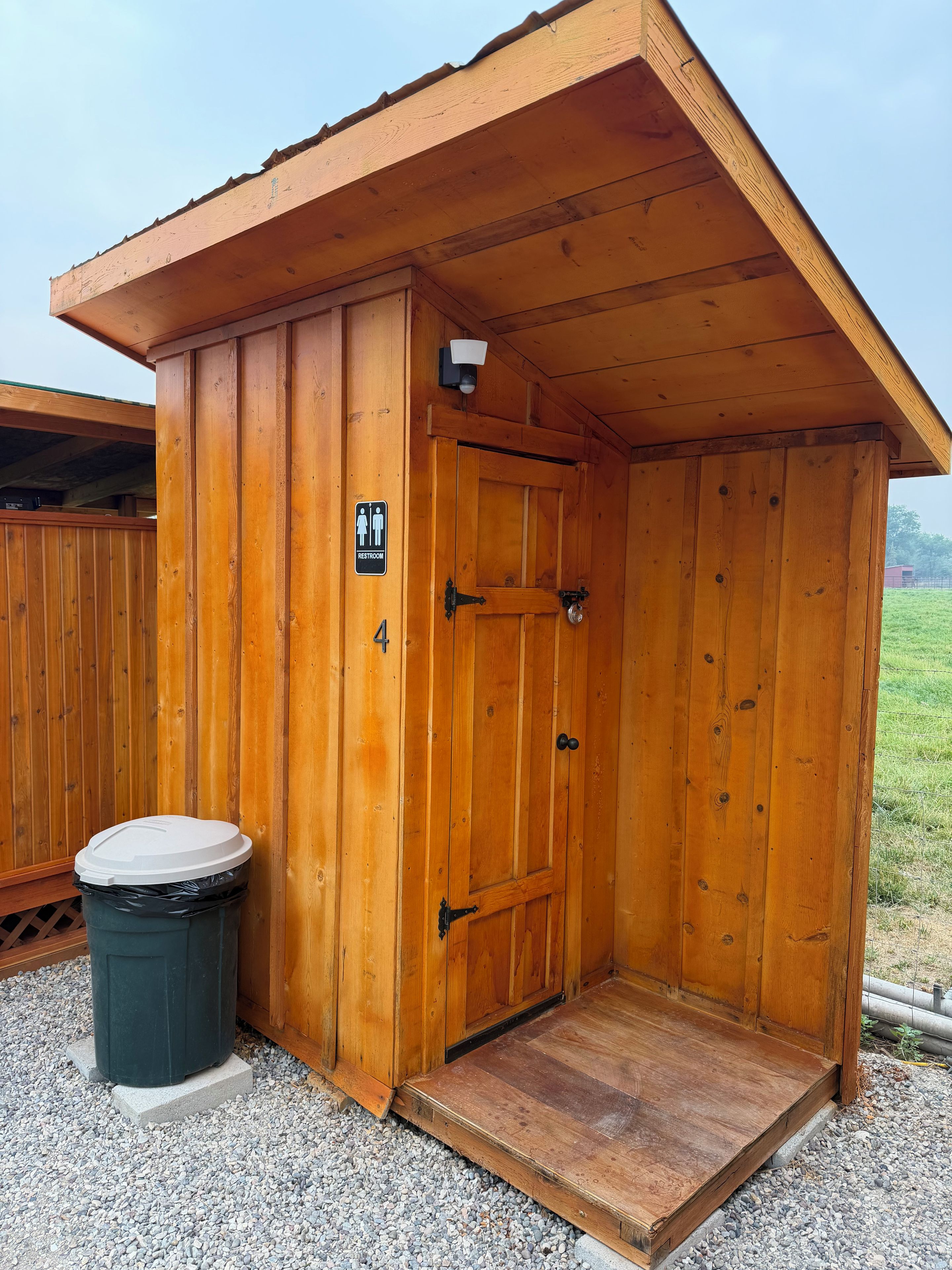 A wooden restroom with a sign indicating it is for men and women, a trash bin, and a door with a handle.