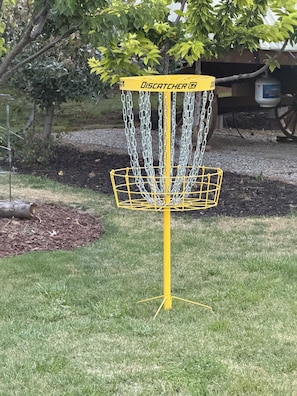 Un panier de disc golf jaune, muni de chaînes et d’un anneau de réception, est installé dans une zone herbeuse, avec des arbres et un bâtiment en arrière-plan.