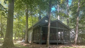 Mökki, jossa on suuri, ikkunallinen kuisti, ja se sijaitsee korkeiden puiden ympäröimällä metsäisellä alueella.