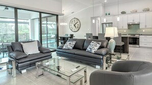 A modern living room with a black leather sofa, a glass coffee table, and a dining area with a chandelier. - VILLA BRILLANTE, A BRAND NEW Luxury Waterfront home  Pool/Spa & Gulf Access (Cape Coral)