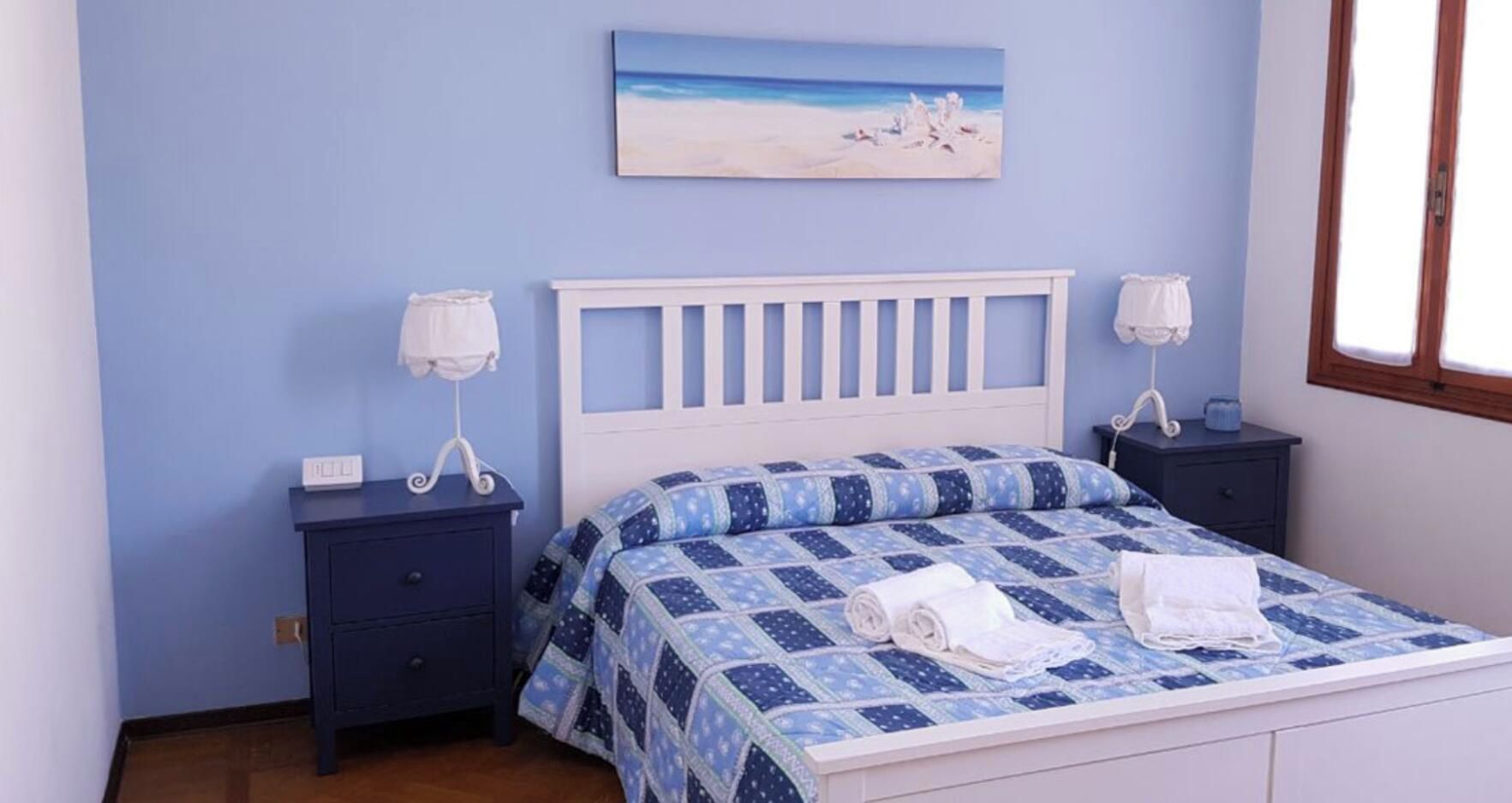 Une chambre avec une couvre-lit à carreaux bleus et blancs, deux tables de chevet avec des lampes, une table de nuit et un tableau représentant une scène de plage accroché au mur.