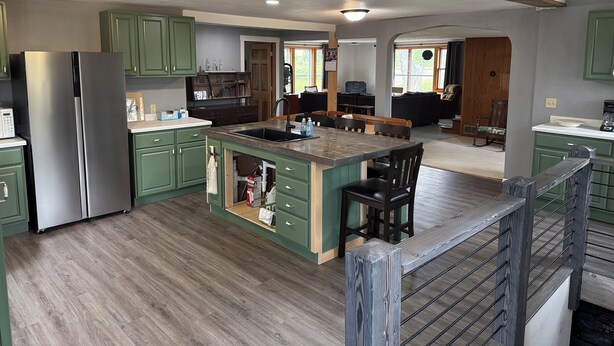 A spacious kitchen with green cabinets, a stainless steel refrigerator, and a wooden island with bar stools. - Rogers Creekside Ranch - Wessington Springs, SD - 3+ Bed, 1 Bath (Wessington Springs)