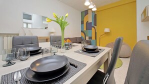 Zona pranzo con tavolo apparecchiato per quattro, con piatti, bicchieri e posate. Sul tavolo c'è un vaso con fiori, e la stanza presenta un design moderno, con una parete gialla a contrasto e un grande specchio.