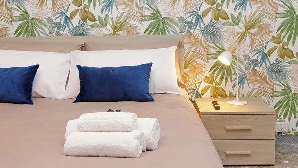 Un letto rifatto con cuscini bianchi e blu, una lampada da comodino e asciugamani piegati su un tavolino di legno.