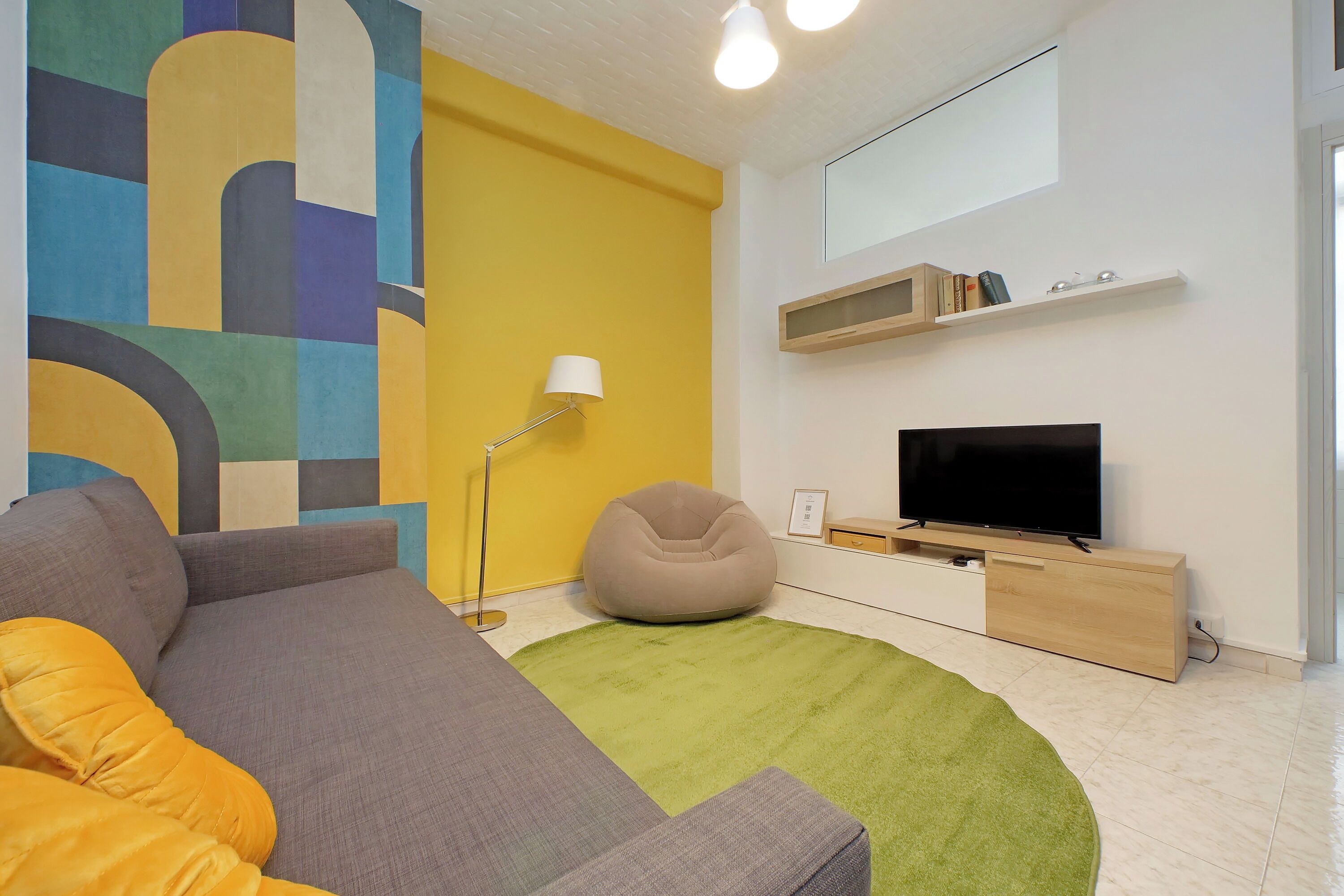 A modern living room with a sofa, a bean bag, a flat-screen TV, a lamp, and a colorful abstract wall.