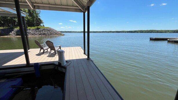 A wooden deck with chairs and a table, overlooking a calm lake with a dock in the background. - Dockside Dreamz- Full duplex lake fun for 12 (Clarksville)