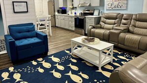 A modern living room with a blue armchair, a brown leather sofa, a white coffee table, and a kitchen area with white cabinets and a stainless steel refrigerator. - Dockside Dreamz- Full duplex lake fun for 12 (Clarksville)