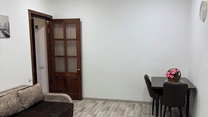 Quarto compacto com sofá, mesa pequena com cadeiras e porta de madeira.