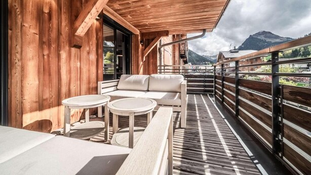 Ein Holzbalkon mit einer Couch und zwei runden Tischen, mit Blick auf eine Berglandschaft.