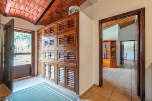 A hallway with a wooden door, a tiled floor, and a view into another room with a bookshelf and a lamp.
