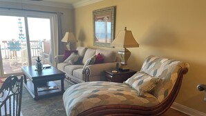 A living room with a sofa, armchair, coffee table, and a lamp. There is a dining area with chairs and a table. A large window with curtains is visible. - Beach Front Townhome – 2 BR, 3 BA - Deck Overlooking Gulf & Steps to the Beach (Panama City Beach)