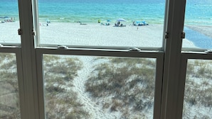 A view through a window with blinds, overlooking a beach with people and beach chairs. - Beach Front Townhome – 2 BR, 3 BA - Deck Overlooking Gulf & Steps to the Beach (Panama City Beach)
