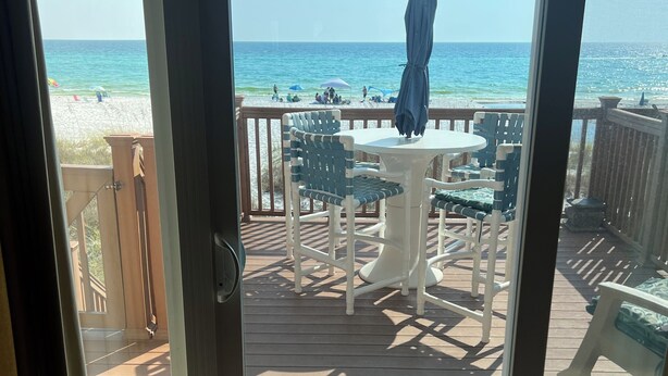 A deck with a table and chairs, overlooking a beach with umbrellas and people. - Beach Front Townhome – 2 BR, 3 BA - Deck Overlooking Gulf & Steps to the Beach (Panama City Beach)