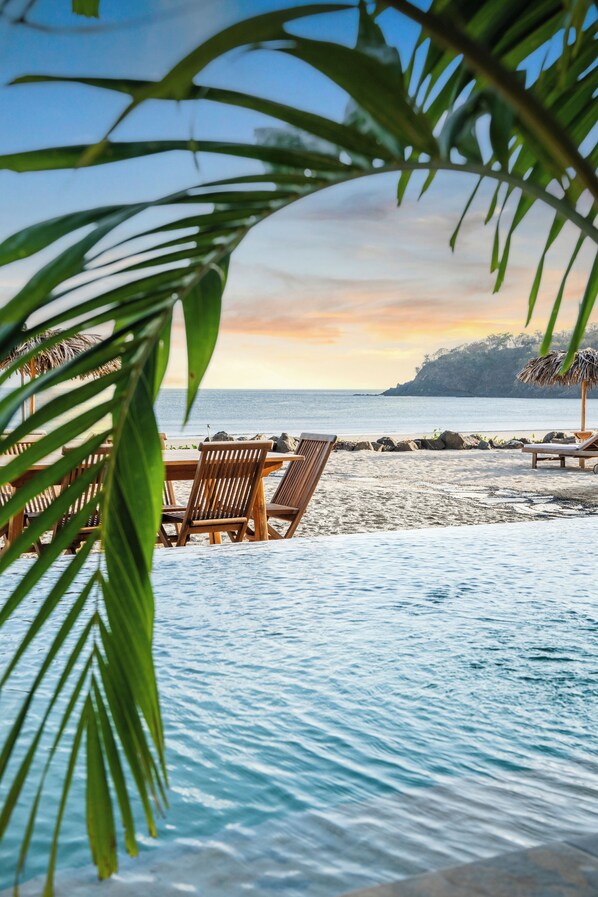 A beachside setting with a wooden table and chairs, a pool, and a view of the ocean and a distant mountain.