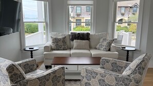 Ein Wohnzimmer mit einer Couch, zwei Sesseln, einem Couchtisch und einem an der Wand befestigten Fernseher. Fenster bieten Blick auf Gebäude und Grünflächen.