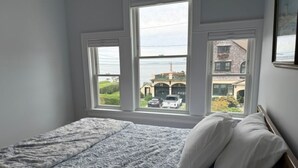Ein Schlafzimmer mit Bett, einem Fenster mit Blick auf ein Gebäude und parkende Autos sowie einem gerahmten Bild an der Wand.