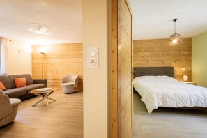 Moderni huone, jossa on puulattia, valkoisilla vuodevaatteilla varustettu sänky, harmaa sohva, pieni pyöreä pöytä ja tuoli.