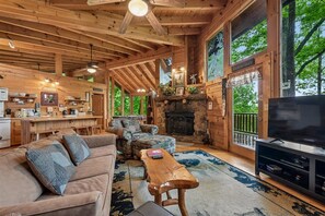 A log cabin interior with a stone fireplace, a television, and a large window overlooking greenery. - Resort Amenities and Game Room Fun Await! (Sevierville)