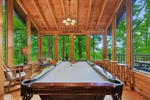 A pool table in a wooden room with large windows, a hanging light fixture, and a sign above the table.