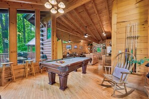 A wooden cabin interior with a pool table, seating area, and large windows overlooking a forest.