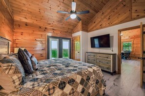 Un dormitorio con techo de madera, una cama con ropa de cama a cuadros, un aparador, un televisor y una puerta al balcón.